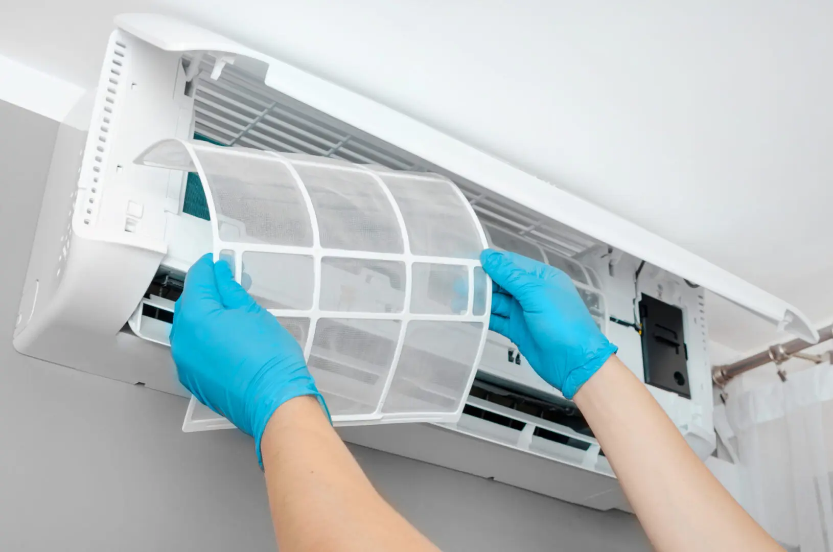 Person wearing blue gloves removes a filter from a wall-mounted air conditioning unit for cleaning.