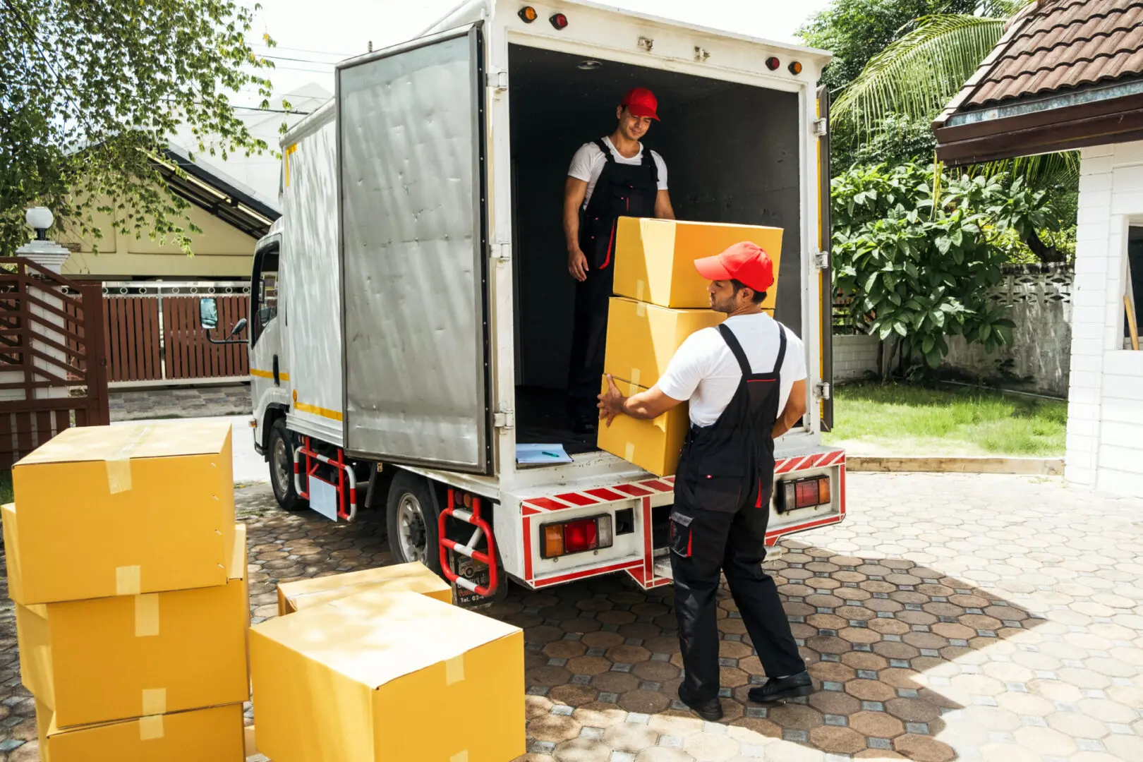 Two workers in uniforms unload cardboard boxes from a truck in a residential driveway. One worker stands inside the truck, and the other carries a box outside.